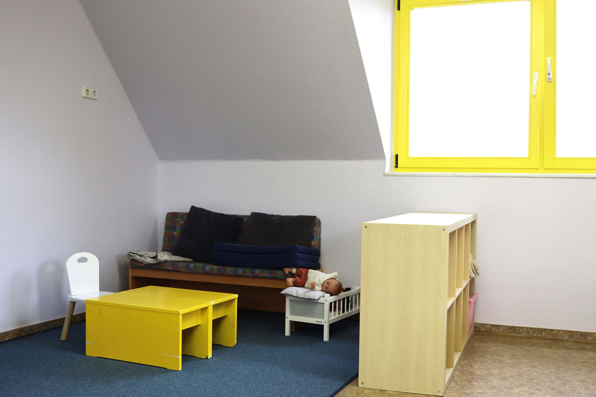 Spielzimmer. Blauer Teppich unter einer Dachschräge. An der Wand ein kleines Sofa mit Kissen, ein kleiner gelber Tisch steht davor, an dem Tisch ein kleiner weißer Stuhl. Neben dem Sofa ein  Puppenbett mit Babypuppe. Als Abgrenzung ein Regal mit Fächern.