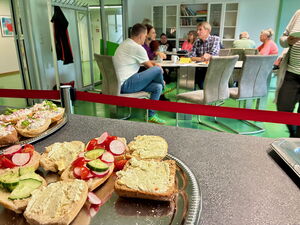 Belegte Brötchen im Vordergrund. Hinten im Bild, Tische, Stühle, lockere Gesprächsrunden.
