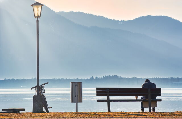 Eine Person sitzt einsam auf einer Bank am Wasser. Links neben der Bank ein Mülleimer noch weiter links lehnt ein Fahrrad an einer Laterne.