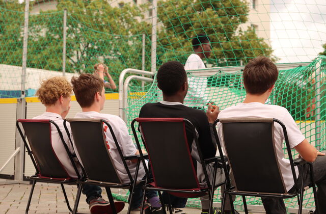 Vier Jugendliche sitzen auf Klappstühlen und sehen dem Fußballspiel zu.