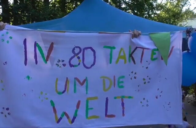 Großes Stofflaken bunt beschrieben, hängt an einer Wäscheleine. Text darauf: "In 80 Takten um die Welt"