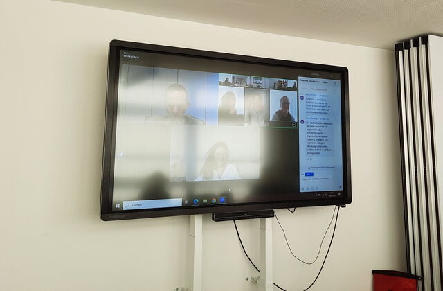 Monitor an der Wand. Ein Online Meeting. Mehrere Menschen sind zugeschaltet.