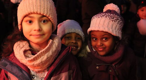 Drei Mädchen. Mit Winterkleidung, Mützen, Schal, Winterjacke. Im Alter von ca. 5 - 7 Jahren. Zwei der Mädchen sind afroafrikanisch (People of Color). Alle drei lächeln fröhlich.