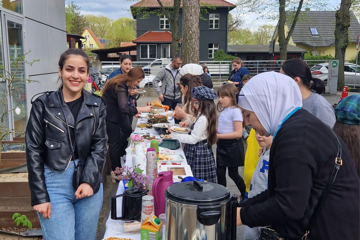 Draußen, ein Büfett, langer Tisch mit Getränken und Speisen. Kinder und Frauen holen sich was zu essen.