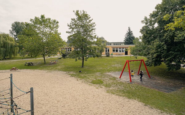 Großes Gelände. Wiese, Bäume, großer Sandkasten. Kinder auf einer roten Schaukel. Spielplatz
