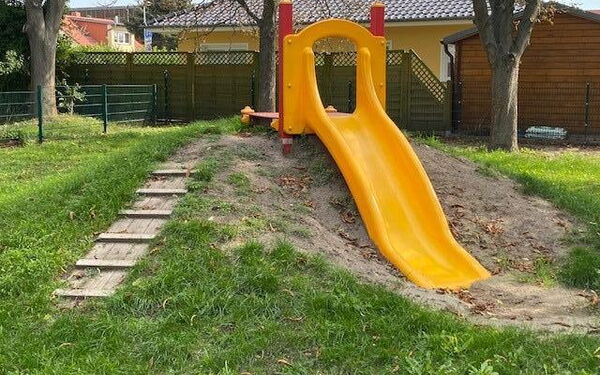 Eine gelbe Rutsche auf eine kleinen Hügel. Wiese, drei Bäume.