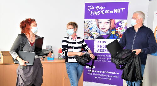 Im Büro Kinderarmut: 2 Frauen und ein Mann stehen mit Corona Abstand und OP-Masken nebeneinander. Sie halten Laptops in den Händen. Ein Frau hält gleich zwei Laptops.