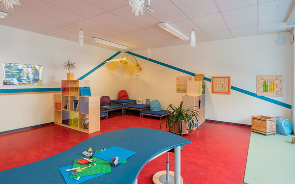 Großes Zimmer mit Sitzecke, zwei offene Regalen auf den Boden gestellt trennen die Sitzecke etwas vom Raum ab. Ein geschwungener blauer Tisch mit einem Hocker befindet sich etwas Abseits der Sitzecke.