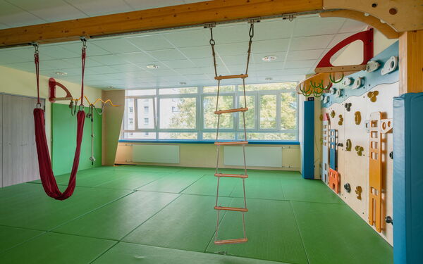Sportraum. Hängeleiter an einem Holzbalken. Der Fußboden ist mit grünen Matten ausgelegt, An der Wand viel Griffe, Reifen und Tritte zum Klettern.