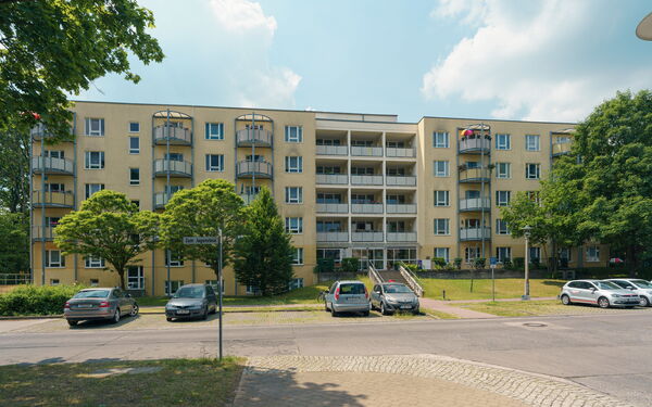 Eine Straße, wenige parkender Autos. Dahinter und großes Wohnhaus mit gelber Fassade und Balkonen.