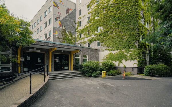 Eingang des Gebäudekomplex. Das Gebäude ist mit Efeu bewachsen. Zum Eingang Stufen und links eine geschwungene Rampe.