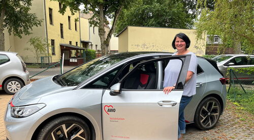 Ein silbernes VW Auto mit dem Logo der AWO Sozialstation auf der Fahrertür. Die Tür ist geöffnet und eine Frau mit schwarzen vollen Haaren, einem hellen T-Shirt und blauer dreiviertel-Jeans steht an der Fahrertür und und lächelt in die Kamera.
