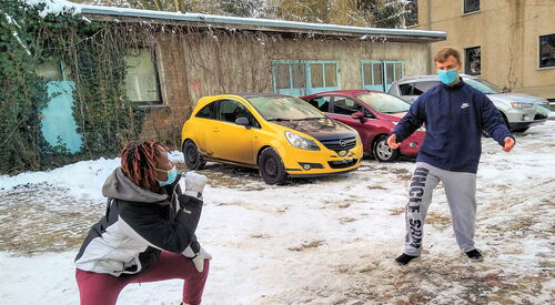 Tanzszene im Freien. Es ist Winter, Schnee liegt, parkende Autos. Eine schwarze Tänzerin mit OP Maske ist in einer breitbeinigen Hocke einem Tänzer mit OP Maske zugewandt. Der weiße Tänzer hat die Arme ausgebreitet. Sie stehen etwas von einander entfernt.