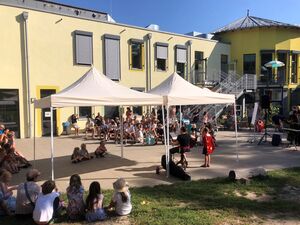 Draußen. Schulgebäude gelb. Auf dem Platz davor zwei Pavillons. Kinder und Eltern sitzen auf Stühlen und auf dem Rasen und schauen dem Kulturprogramm zu.