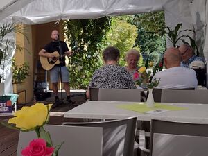 In einem großen weißen Veranstaltungszelt. Musiker mit Gitarre und Mikrofonständer. Davor ein Schild mit seinem Namen, Robert Bernier. In dem Zelt Tische mit Vasen mit roten und gelben Rosen. An einem Tisch sitzen 4 alte Personen, 2 Frauen, 2 Männer