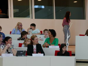 Kinder und Erwachsene sitzen im Plenarsaal. Einige Erwachsene haben Namensschilder auf den Tischen vor sich.