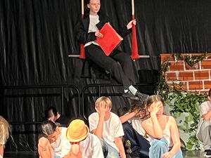 Bühnenszene: auf eine Schaukel sitzt eine Person/Puppe in schwarz-weißer Kleidung. Darunter Kinder in der Hocke die die Hand vors Gesicht legen.