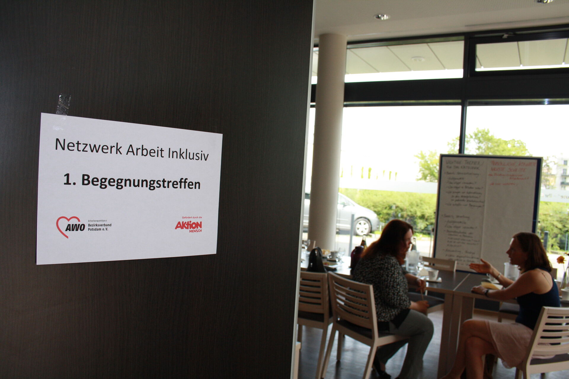 Blick in einen Raum. Zwei Frauen unterhalten sich angeregt. Sie sitzen an einem Tisch.
