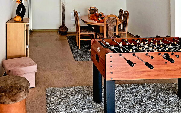 Tischkicker aus dunklen Holz auf grauem Teppich. Im Hintergrund ein ovaler Tisch mit 6 Stühlen steht auf auf einem grauen Teppich.