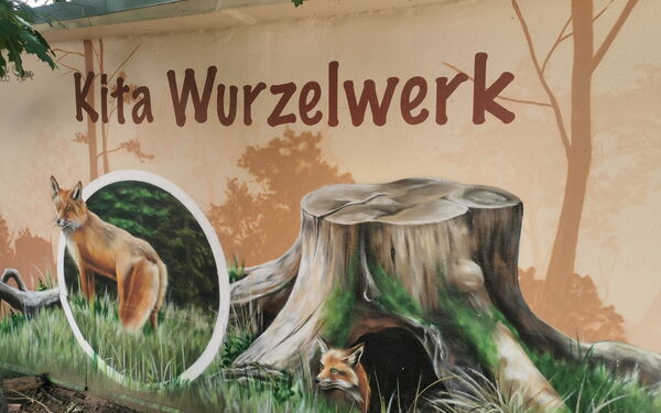 Eine Häuserwand mit einem Wandbild von eine Fuchsbau unter einem großen Baumstumpf. Ein Fuchs schaut aus dem Bau. Drüber steht groß: Kita Wurzelwerk