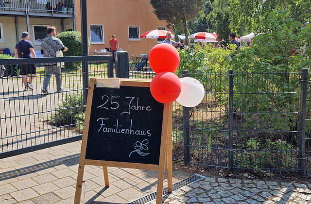 Draußen, Sonnescheint, eine Klapp-Tafel steht am Eingang geschmückt mit roten und weißen Luftballons. Auf der Tafel steht mit weißer Kreide geschrieben: "25 Jahre Familienhaus"