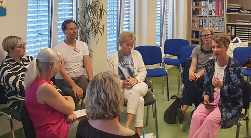 Im Kreis angeordnete blaue und graue Stühle. Menschen sitzen im Kreis. In der Mitte Sitzt eine Frau mit rosa Stoffhose, weißem T-Shirt und blauer Stickjacke und spricht zu den 5 Teilnehmenden.