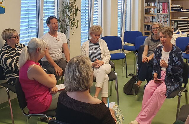 Im Kreis angeordnete blaue und graue Stühle. Menschen sitzen im Kreis. In der Mitte Sitzt eine Frau mit rosa Stoffhose, weißem T-Shirt und blauer Stickjacke und spricht zu den 5 Teilnehmenden.