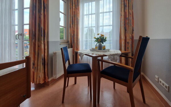 Zimmer, zwei Stühle blau gepolstert und kleiner Tisch vor Fenstern.