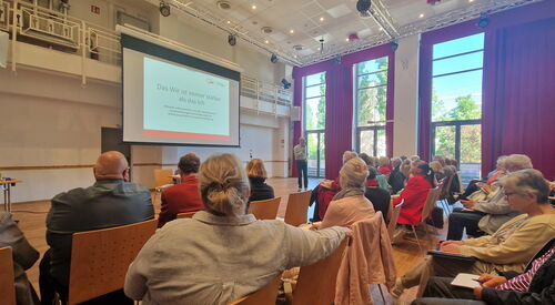 Großer Saal. Viele Stuhlreihen wo Menschen sitzen, Leinwand forne, ein Redner mit Mikrofon steht vorn.