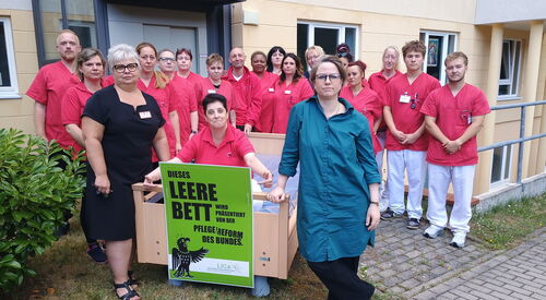 Viele Pflegende in Arbeitskleidung vor einer Einrichtung. Ein Pflegebett steht vor ihnen, darin sitzt eine Pflegerin und zeigt auf ein angebrachtes grünes Schild.