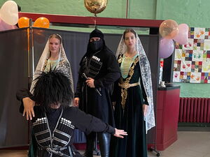 Vier Personen in schwarzen Kostümen mit weißen Schleiern und Verzierungen, eine davon kniet mit ausgebreiteten Armen im Vordergrund.