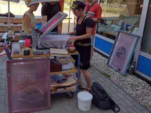 Live Siebdruck mit der Siebdruckwerkstatt. Eine Frau in dunkler kurzer Kleidung legt ein T-Shirt in die Siebdruckmaschine. 3 weitere Menschen sehen sich das an.