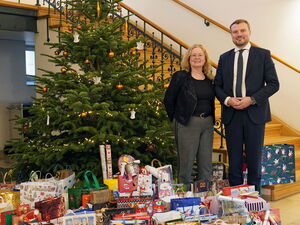 In einem großen Flur mit Treppe und geschmückten Tannenbaum. Neben dem Baum eine Frau und ein Mann in Anzug und Krawatte. Vor dem Tannenbaum sehr viele große und kleine Geschenktüten.
