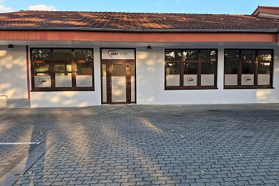 gepflasterte Parkfläche mit grauen Steinen, im Hintergrund ein Gebäude mit einer weißen Fassade, einem roten Dach, mit einer Glastür mittig, links von der Glastür eine Fensterglasfront, rechts der Glastür zwei Fensterglasfronten