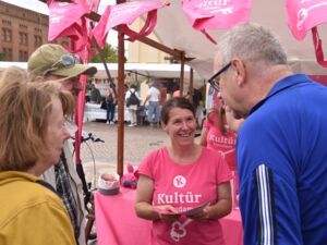 Frau verteilt Kultür Flyer an Interessierte. Frau trägt pinkes Kultür Potsdam T-Shirt und brauen Haaren. Sie unterhält sich mit 3 Menschen die am pinken Kultür-Stand stehen.