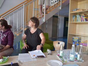 Zwei Frauen sitzen am Tisch im Raum. Auf dem Tisch Wasserflaschen, Gläser. Eine Frau hat Unterlagen vor sich auf dem Tisch liegen und greift danach.