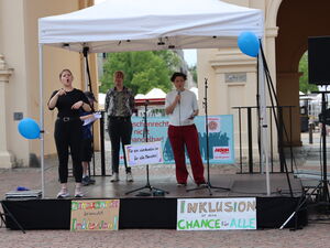 Eine Bühne mit Gebärdendolmetschung und einer Frau mit Mikrofon. Dabor Protestplakate: Demokratie braucht Inklusion.