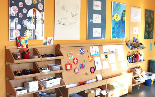 Bastelzimmer. An der Wand große Bilderrahmen mit verschiedenen kleinen Bildern und Zeichnungen. Darunter eine Art Bastel-Regal mit mehreren Ablageflächen für Scheren, Stifte, Papier.