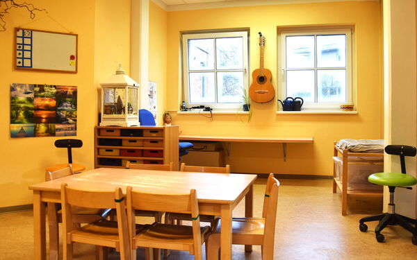 Bastelzimmer. Tisch mit 6 Stühlen. Gelbe Wände. Zwischen zwei Fenstern hängt eine Gitarre an der Wand.