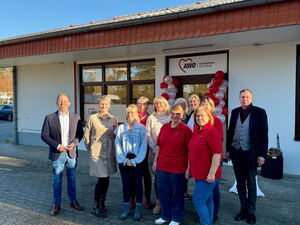 11 Personen, zwei Männer links und rechts außen stehen vor einem weißen Gebäude. Der Eingang ist mit roten und weisen Luftballons geschmückt.