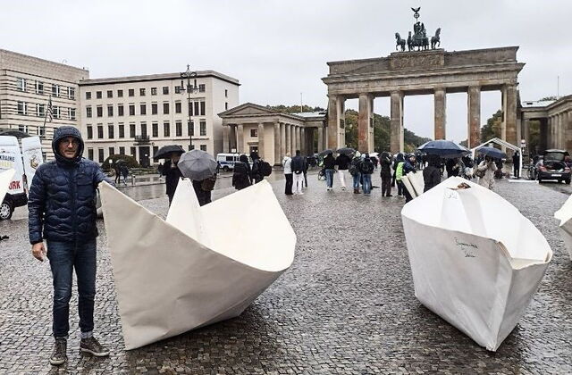 Vorplatz des Brandenburger Tor in Berlin. Regenwetter. Menschen mit Regenschirmen und Kapuzenjacken. 2 XXL-Origami-Papierboote stehen auf dem Platz. Ein Mann steht neben einem der Boote, er trägt eine dunkelblaue Regenjacke die Kapuze hat er aufgesetzt.