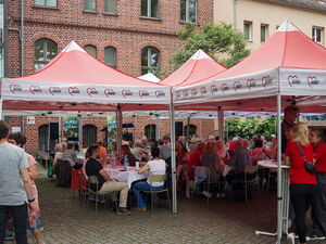 Einen Innenhof mit roten AWO Pavillons mit Tischen und Stühlen wo viele Menschen zusammen sitzen, essen und trinken.
