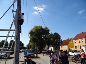 Draußen, blauer Himmel. Eine Kletterwand ist aufgebaut. Ein Kind klettert hinauf und wird durch ein Seil gesichert.