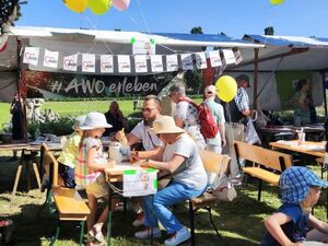 Vor Marktständen sind Tische und Bänke aufgestellt, an denen 2 Kinder und 2 Erwachsene sitzen und die Kinder malen Bilder. Der Stand ist mit Luftballons und Wimpeln geschmückt. Die Sonne scheint.