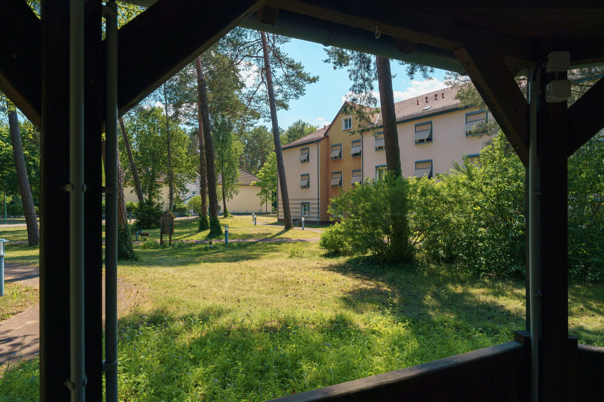Blick aus einem Holzpavillon auf Wiese, Bäume und ein Wohnhaus.