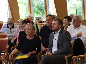 Mehrere Menschen sitzen in einem großen Raum. In der ersten Reihe, eine blonde Frau mit Brille auf den Kopf geschoben, die hat einen gelben Hefter auf dem Schoß. Daneben ein Mann mit weißen Hemd und grauen Anzug.