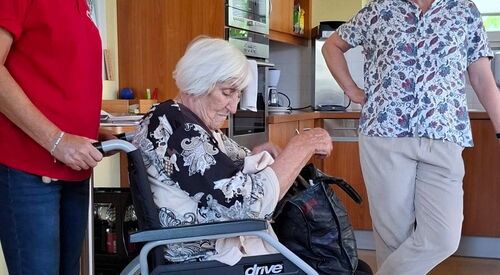 Pflegerin im roten Shirt schiebt alte Frau im Rollstuhl in eine Küche.