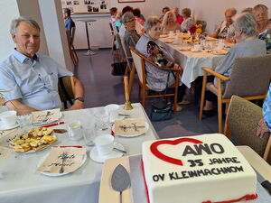 Kaffeetafeln an denen größtenteils ältere Menschen sitzen. Im Vordergrund eine große weiße Torte mit AWO Logo und Schrift.