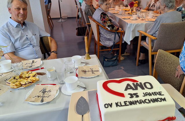 Kaffeetafeln an denen größtenteils ältere Menschen sitzen. Im Vordergrund eine große weiße Torte mit AWO Logo und Schrift.