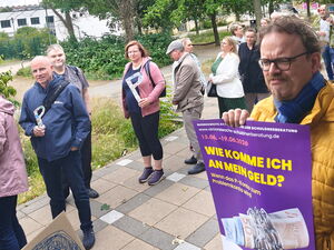 Draußen: Menschen-Schlange. Einige Personen halten ein großes P in der Hand.   Rechts im Bild Mann mit gelber Jacke hält lila Plakat vor sich.